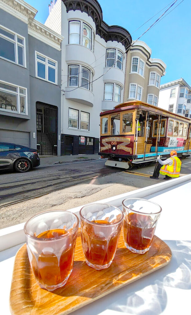 three glasses are on a tray. They have a brown liquid in them, coffee, each mostly full. Behind the coffee tasters is a man pulling a bar on the road to stop the oncoming cable car in the san francisco streets.