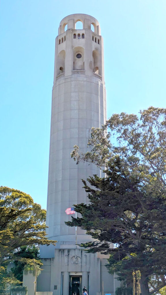 The not so beautiful concrete coit tower in the sky. There are some green trees lining the bottom of it. And an American flag blowing in the wind at the base.