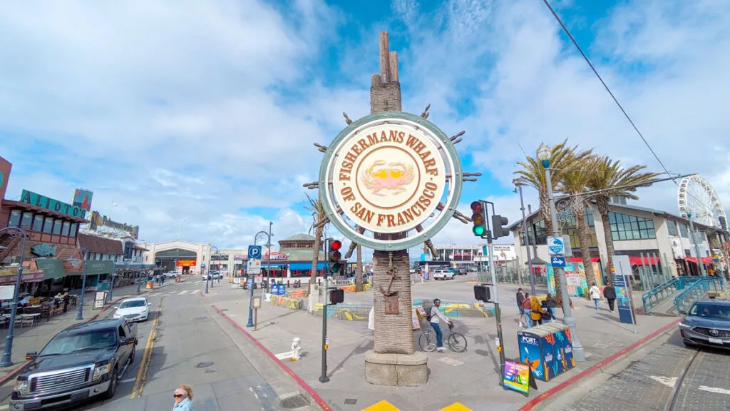 a street corner has a large sign, shaped like the wheel of a ship. In the middle it says in a circle, Fisherman's Wharf of San Francisco.