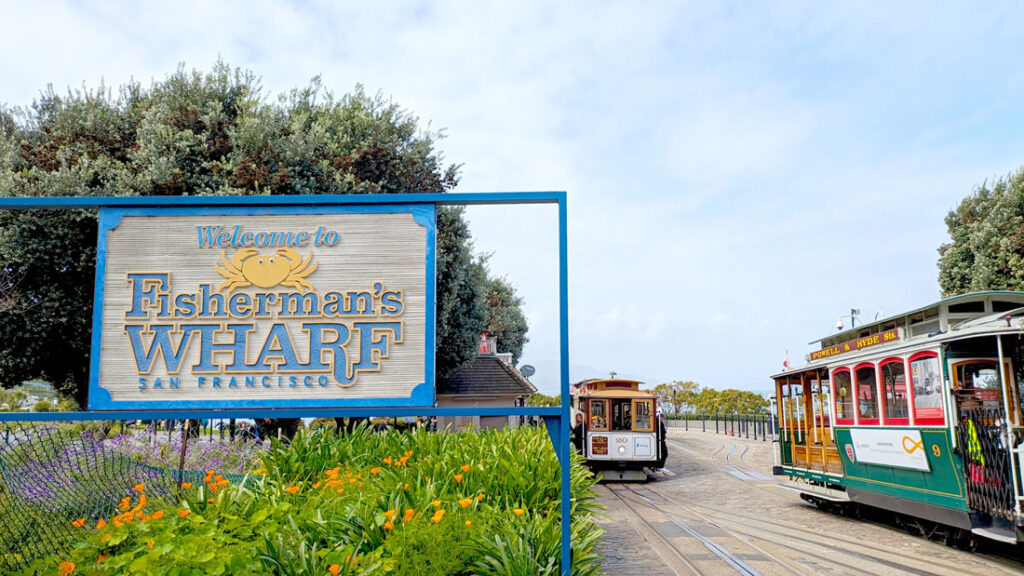 a large old school sign for Fisherman's Wharf San Francisco stands on the left side in some bushes at the base. On the right you can see some tracks, where there's a red and yellow nad a green and yellow cable car on either one.