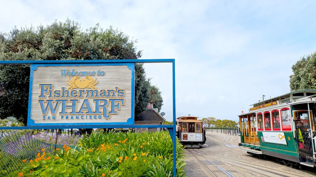 a large old school sign for Fisherman's Wharf San Francisco stands on the left side in some bushes at the base. On the right you can see some tracks, where there's a red and yellow nad a green and yellow cable car on either one.
