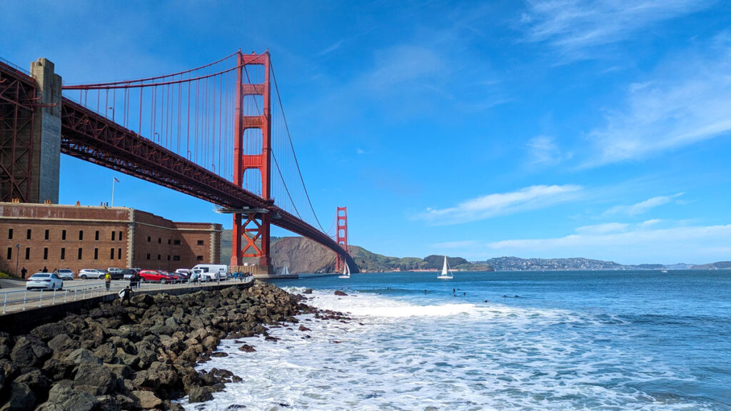 a view of fort point old warehouse building. The ocean waves could hit the building but a rock wall breaks it. It adds a white tips to the blue water that leads underneath the golden gate bridge high above. You can see it go all across the water all the way to the other side.