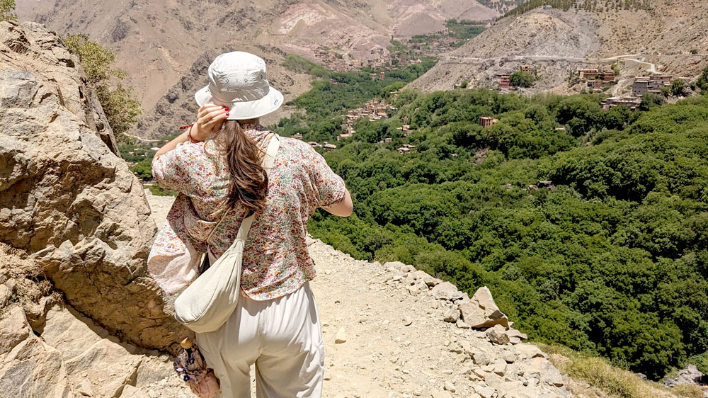 a woman is photographed from behind as she walked on a narrowish path in the high atlas mountains. You can see a small Berber Village across a valley of trees on the opposite mountain. As she walks down, she holds on to the back of her hat so it doesn't fly away into the valley below.