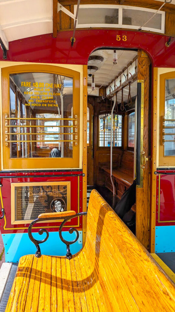 the view from inside a cable car. The sun is shining on a bench facing outward, casting a beautiful rich yellow hue. There's a burgundy wall with a door behind it, separating the inside booth. On the window you can slightly make out the yellow print where it says single fare $8.00, one way, no transfers