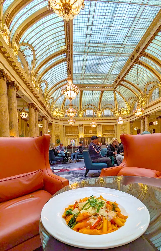 a bowl of pasta with tomato sauce and spinach sits on a glass table. The glass table reflects the ceiling above it, a jaw dropping gold and glass dome with delicate chandeliers hanging from the top. There are two empty red armchairs at the table, they look very cozy to sit in . Behind the chairs the rest of the space is packed with people sitting at similar tables enjoying some snacks and drinks. This is one of the most unique San Francisco experiences!