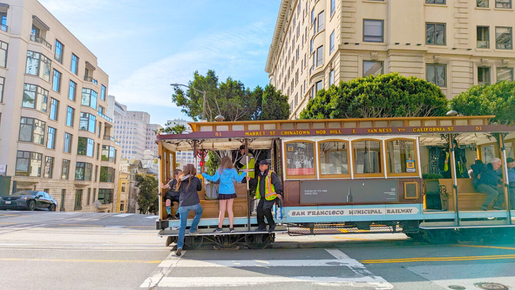 a cable car is stopped in the middle of an intersection on the streets of san francsico. Two people are hanging off the sides ,while a woman steps on to the car. It's not a full car, no one is even sitting on the benches, but they are opting to hang out on the rails instead.