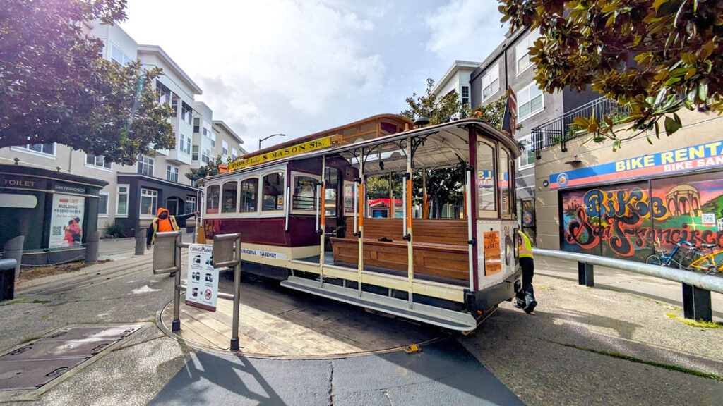a cable car on a circular wood lazy suzan in the middle of a paved road. Two employees are on either end, manually turning the powell mason cable car around in san francisco
