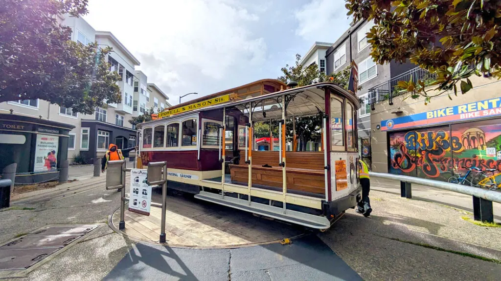 a cable car on a circular wood lazy suzan in the middle of a paved road. Two employees are on either end, manually turning the powell mason cable car around in san francisco