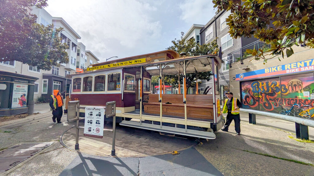 two cable car drivers are in the middle of turning the Powell & Mason cable car around. It's on a wooden turn style on the ground.