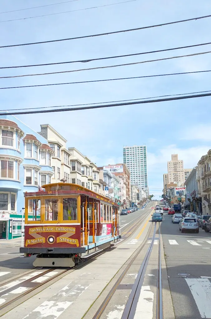 the view from van ness avenue all down california street, you can see the cable car lines down the entire street, cars are driving over them. There's an approaching red and yellow san francisco cable car. beautiful victorian homes line the street on eitehr side.