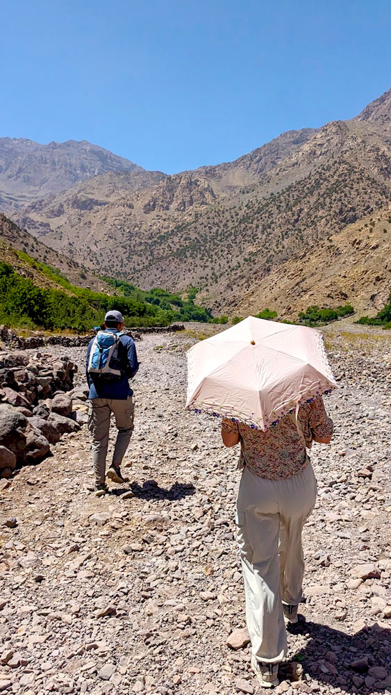 a woman and a man walk through a valley in the high atlas mountains on a gravel path. The man is fully covered, with a backpack. The woman wears a Tshirt, but has a sun umbrella to shade her from the beating sun. There isn't a cloud or tree for shade,