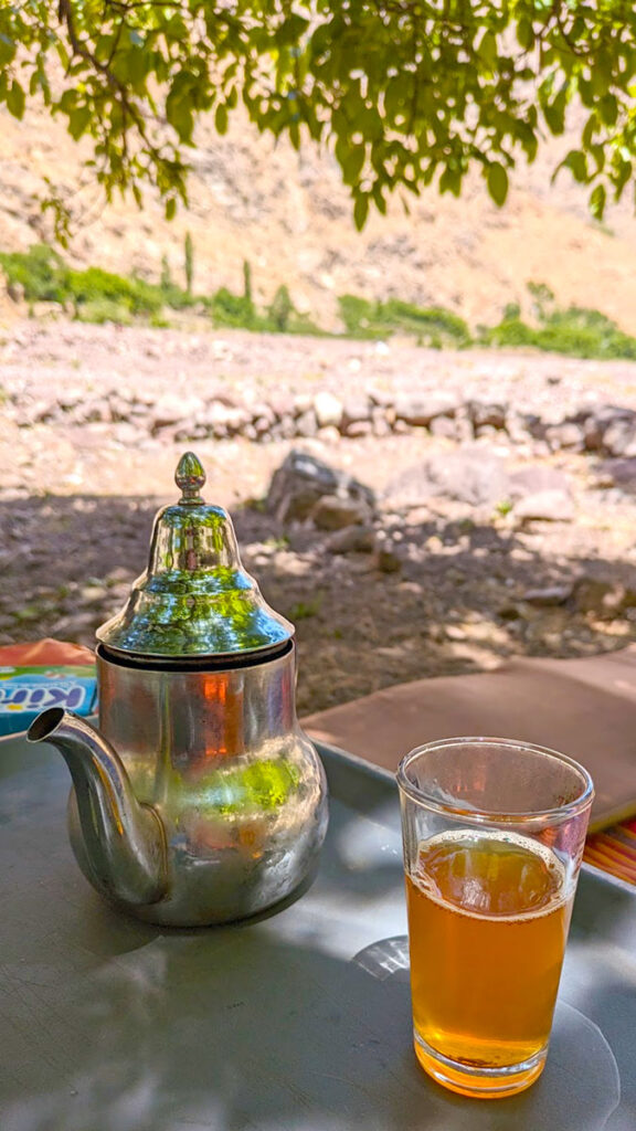 The best experience on a hike in the high atlas mountains. A silver moroccan teapot sits on a table with a glass full of mint tea next to it. The tea is being enjoyed outside on the middle of the mountains, under a tree for shade.