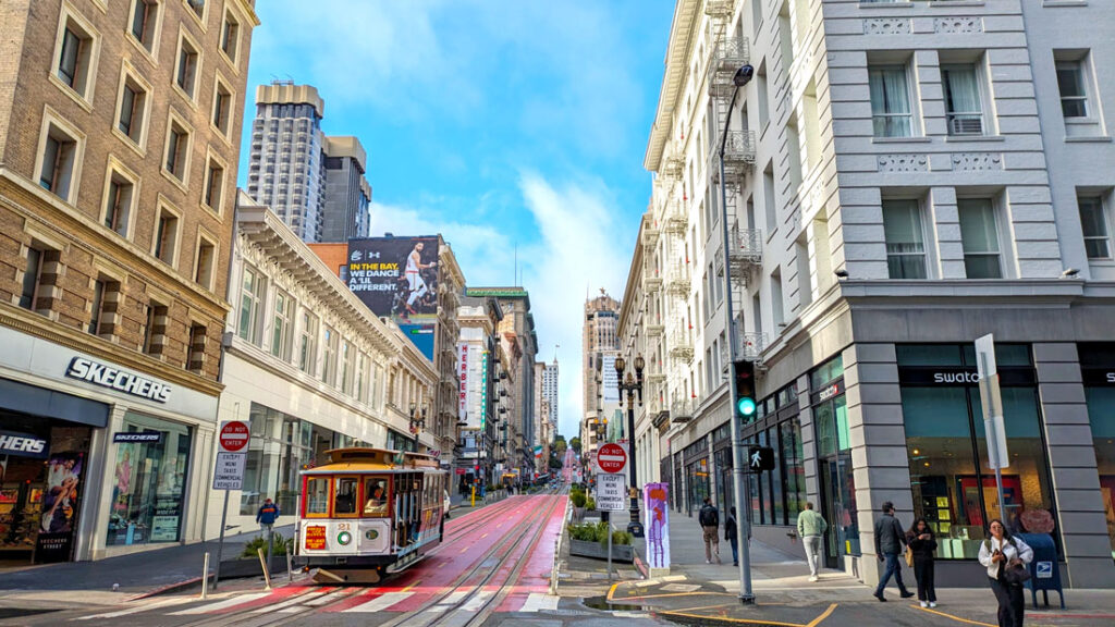 a san francisco cable car at the start of an intersection. Old style buildings are on either side of the street, the sun is hitting the buildings on the right. The street the cable car is on is painted bright red. People are walking on the sidewalks. The sky is bright blue with a few white whispy clouds.