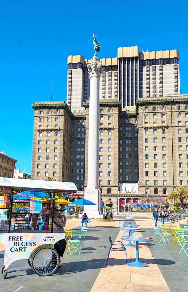 a large courtyard that's far from empty. Colourful blue, yellow, and green tables and chairs fill the same. There's a free Recess Cart that has games for people to play. There's a tall monument directly in the middle and at the back is a historic looking large building with brown stone and intricate designs