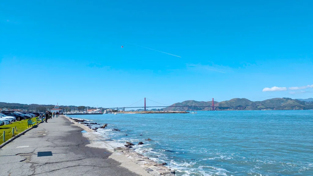 a cracked pavement sidewalk looks like it's leading directly into the bay, but people are still walking on it. There's grass and then a parking lot on the other side of the pavement. In the distance you can make out some boats docked at the port. But the main attraction is just a bit farther, the outline of the slightly reddish golden gate bridge connecting San Franciscos to the other bay.