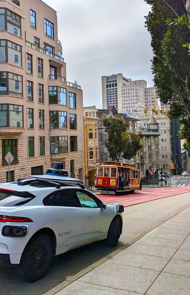 a white car in the foreground. On the back door it says in grey a simple WAYMO. The car definitely has a unique look to it, with extra side mirrors in the front, and a pyramid on the roof. In front of it is on oncoming cable car going up the steep san francisco hill. It's an amazing juxtaposition of new vs old modes of transportation