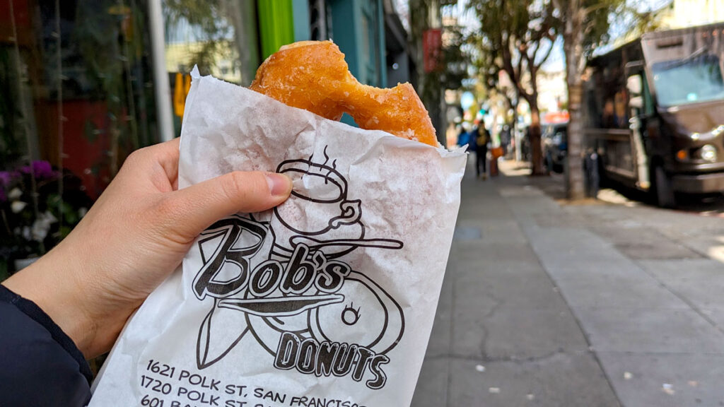 a hand is holding up a white paper bag with a black logo of a donut and a cappuccino cup with Bob's Donuts printed on the front. There's a glazed donut sticking out the top with one bite taken from it (you can't resist these!). Blurred in the background is a sidewalk where you can see some trees and the sunlight shining through.