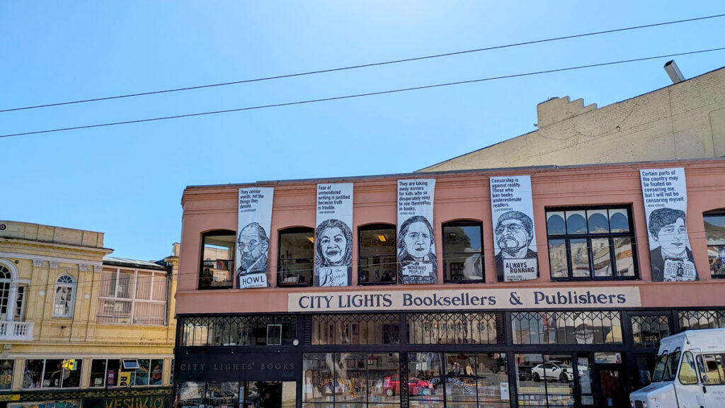 the storefront for city lights booksellers and publishers in a brown brick building. There are tall posters of Beat poertry authors with their famous quote written on them.