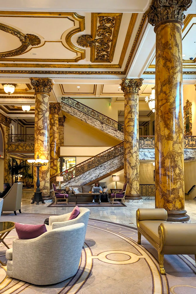 the ornate over the top decorative lobby of the Fairmont San Francisco. A large circular carpet in a brushed purple grey adds a coziness under the grey and purple arm chairs. There are three large brown grained almost gold marble pillars holding up the ceiling. The ceiling is also not left alone, it has gold decals on it. There's also a grand staircase where guests can walk down and feel like royalty.