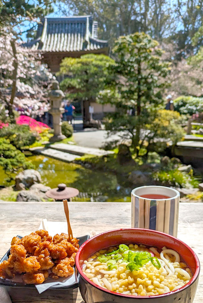 A bowl of ramen, a plate of fried chicken, and a small teacup are on a wood ledge baking in the sunlight. Behind them, slightly blurred is a koi pond, a Japanese style gate, and a lot of green lush trees.