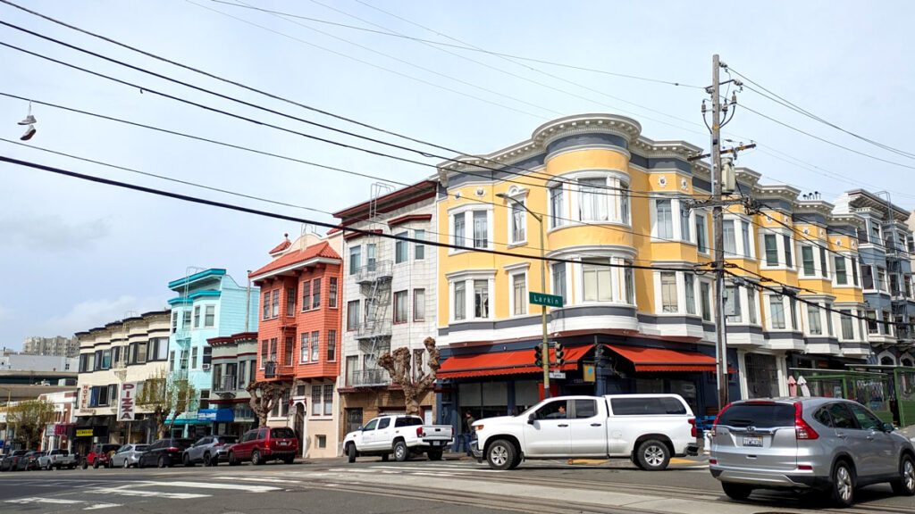 a san francisco street corner filled with unique architecture to admire. The corner building is three stories, with a shop below and bay window columns. The building is painted a bright happy yellow. Next door on it's left is a completely different house, but it's still attached, this one is painted bright blue with a new style, then a red house, then another blue one.