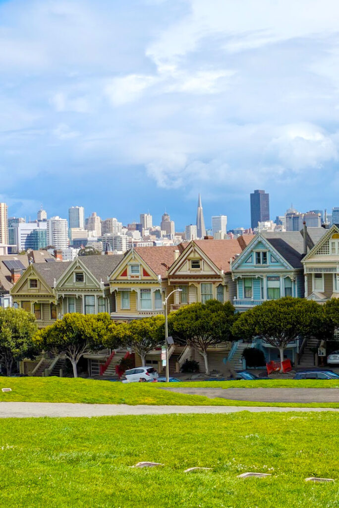 the painted ladies of san francisco across from green grass of alamo square. TShort round green trees line the bottom of the homes. There are six homes in a row, on an incline going up the hill. Each one is a victorian style, and very similar design, but painted a different colour. You can see the cities skyline in the backgorund. Although the homes are sunny, the cloud above look thick and scary.