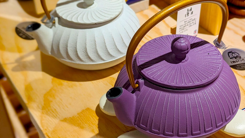 two cast iron teapots in japan, a purple one in the front, and a white one sits behind it.