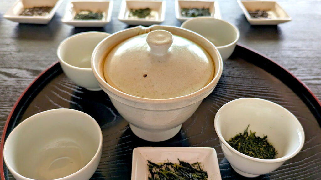 a tay with a tea setting on it, there are four small tea cups, and a traditional tasting japanese teapot. In one of the cups, there are green tea leaves, and there's also a plate next to it with more tea leaves. In the background are 5 more small plates each with their own type of Japanese green tea to try.