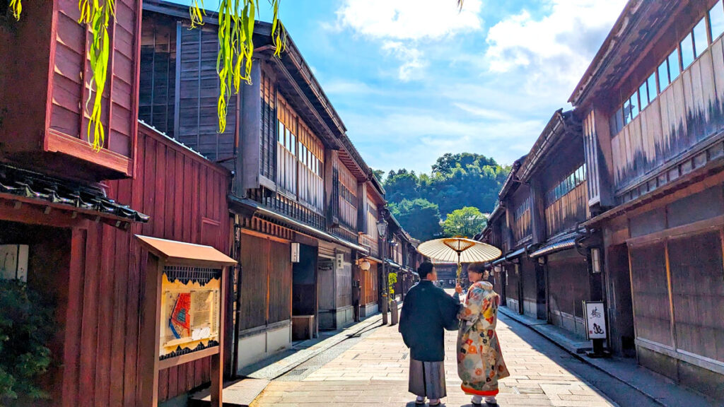 a couple walks away from the camera, they are wearing traditional japanese clothes, with a paper sun umbrella above them. They are walking through what looks like feudal japan, a narrow street, with old wood attached homes lining either side.