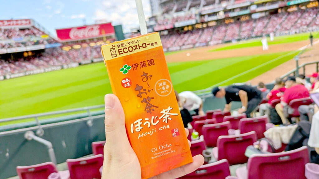a hang is holding up an orange juice box in front of a baseball field and stadium. Except, it's not actually filled with juice but hojicha tea.