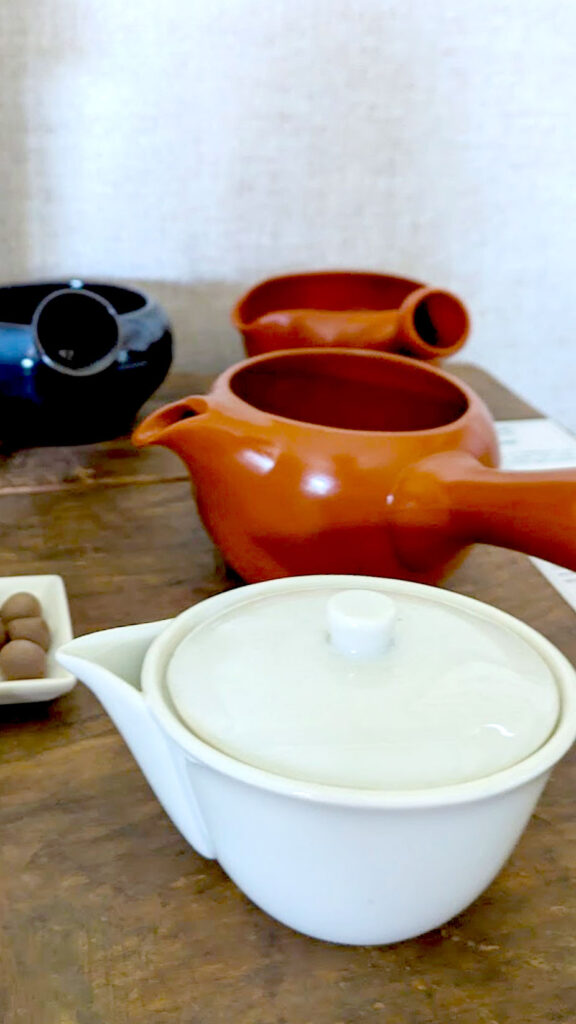 a unique more traditional japanese tea pots for souvenirs. There's a white one in front, with a spout, but no handle. Behind it is a red clay one without a lid, and the handle is on the side instead of behind the spout