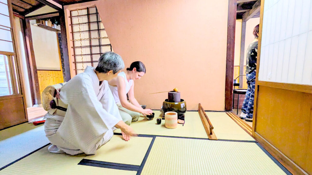 two women kneal on tatami matts next to an odd looking clay pot, a traditional japanese kettle. The farther woman is holding a bowl on the floor, whisking a liquid inside, while the other woman is showing her with her hands what to properly do.