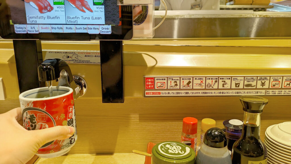 a hand is holding up a large tea cup under a spout, except instead of giving cold water it's filling up the cup with hot water for tea. This is underneath an ipad where you can see pictures of sushi rolls to order.
