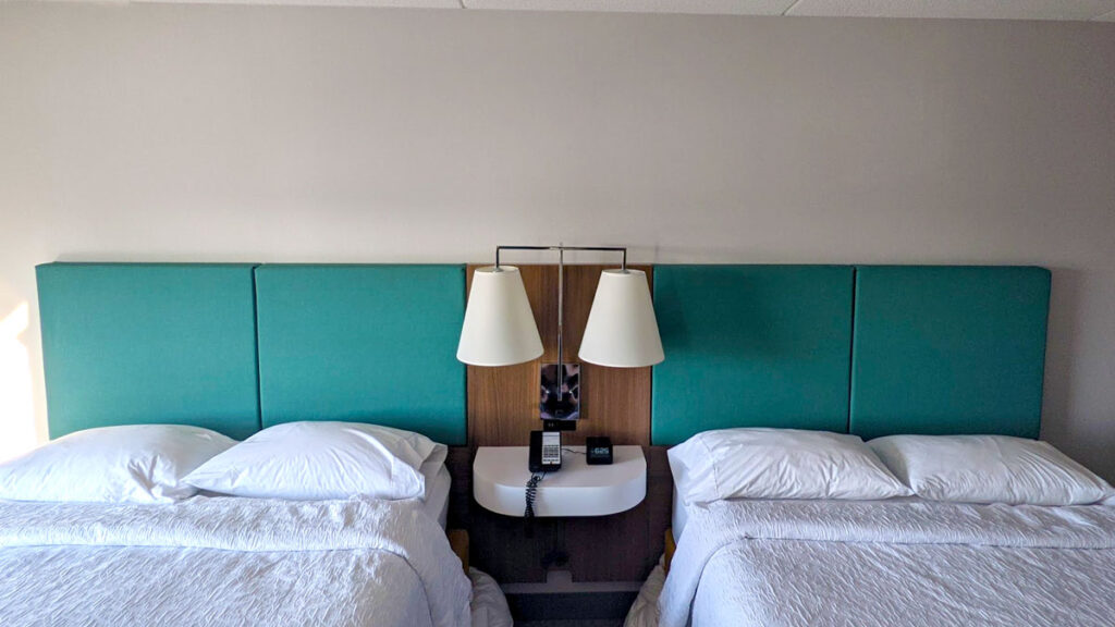 two double beds are side by side on the same wall. There's one bedside table between the two of them with two lights above. The beds are nicely made with white linens, but to add colour to the room and photo, the headboards are teal. 