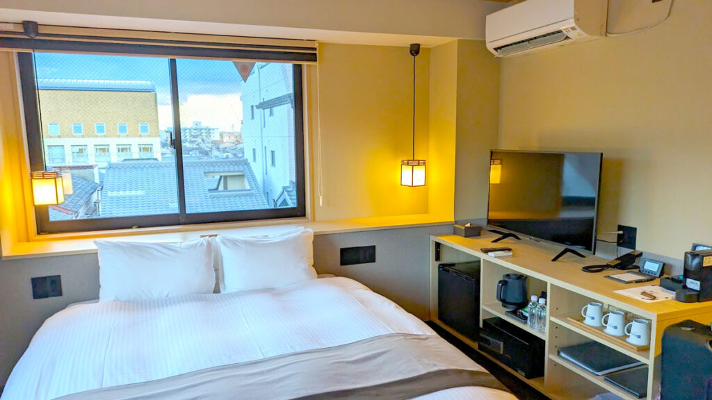 A hotel room in Kyoto, a large bed sits under a window in the middle of the room. On the adjacent wall is a built in cabinet with open shelves offering different hotel amenities. On the top is a TV directly facing the side of the bed. 