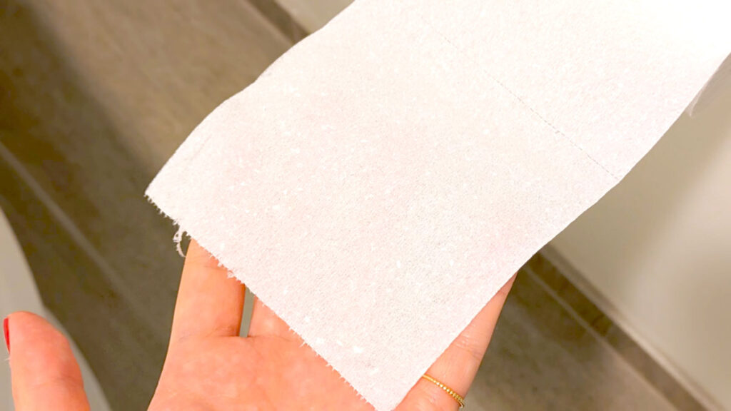 a hand holding out a piece of toilet paper, you can still see her fingers behind the piece of toilet paper, really showing you how see through and thin the hotel toilet paper is. 