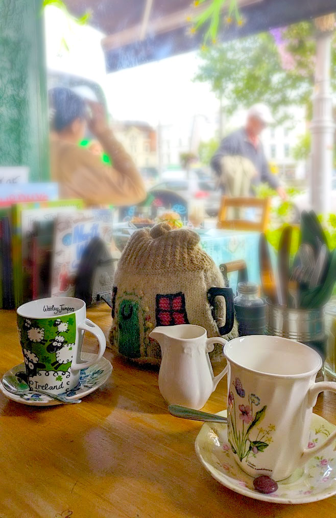 two tea mugs and a tea pot covered in a tea cozy on a wood table by the window. The tea cozy looks like a little house. The mugs are Ireland themed, one is green with sheep on it. There are books on the desk blurred in the background and people outside sitting on the patio and walking by.