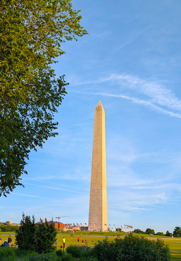 The National Monument is a tall, lean structure rising from the otherwise flat field around it. It's lit up in the golden hour sunlight. Around the base of the monument is a circle of American Flags - in case you forgot where you were. 