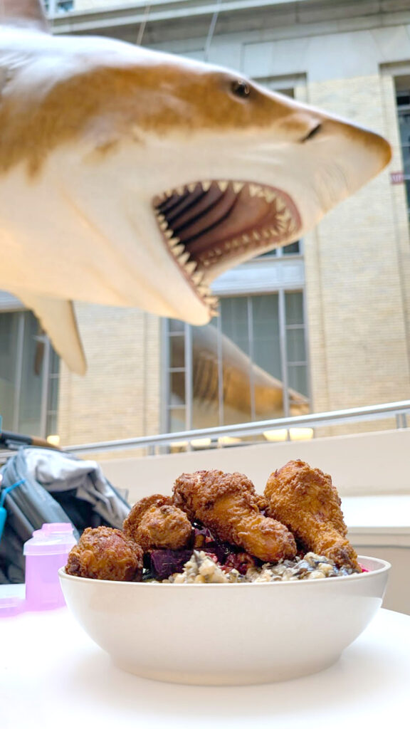a giant shark comes in from the top left corner of the photo, slightly out of focus. The shark's mouth his open, looking like he is wanting to eat the bowl of food on the table below it. It's filled with fried chicken drumsticks that make the bowl overflow on the top. 