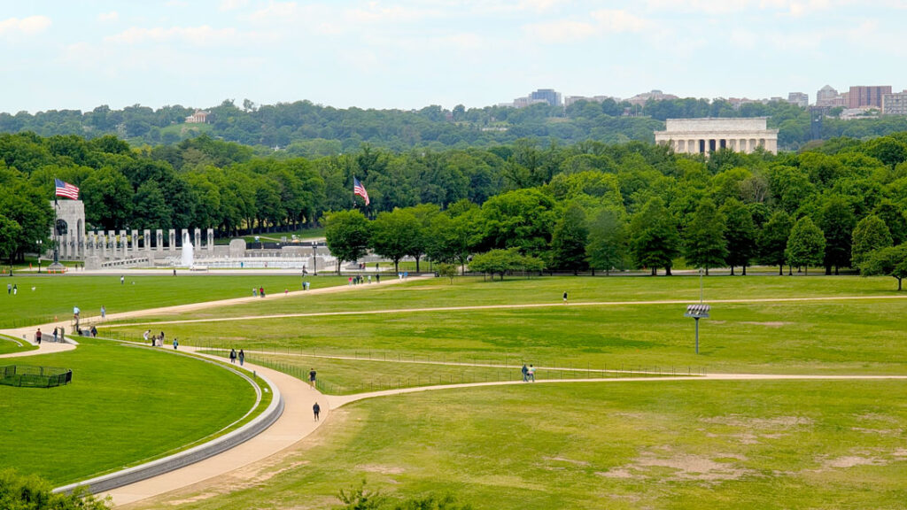 the view of the national mall from above. You can really see how far each monument is from each other. Dots of a few people walk on the allotted path to get to the WWII Memorial. Then are are a forest of trees before you see the top of the Lincoln Memorial poking out from above. The day is overcast adding an ominous hue to the photo. 