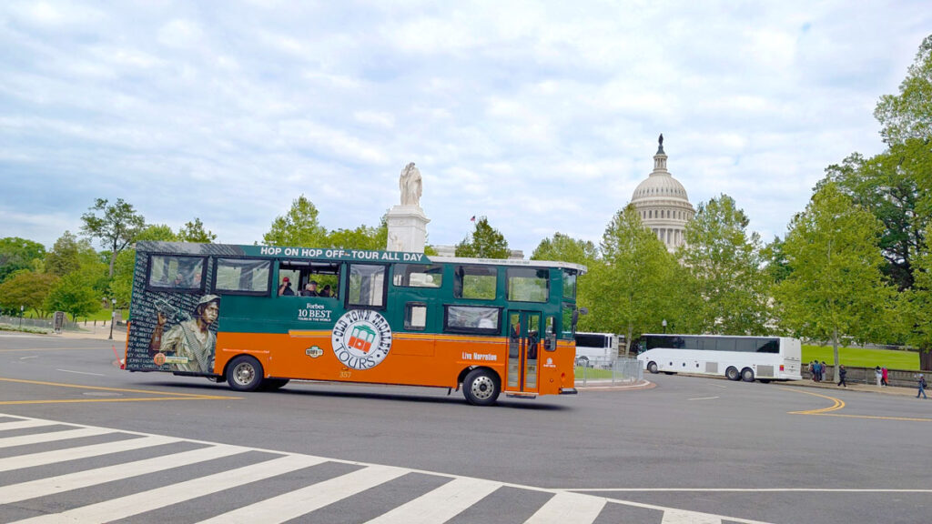 A trolley hop on hop off bus tour drive in front of the u.s. capitol in a roundabout. The buss looks old timey, the bottom is orange, and the top is a darker green. There's one group sitting on the bus with the windows open, they're looking out and taking a photo. 