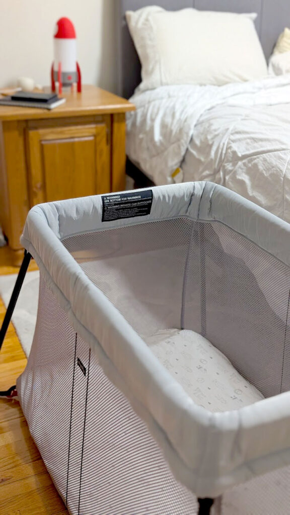 the babybjörn travel crib is the the forefront of the frame, it's light grey frame and completely mesh sides with black legs sticking out. Blurred behind it is a real bed, a wooden bedside table, with a rocket ship light on top of it. 