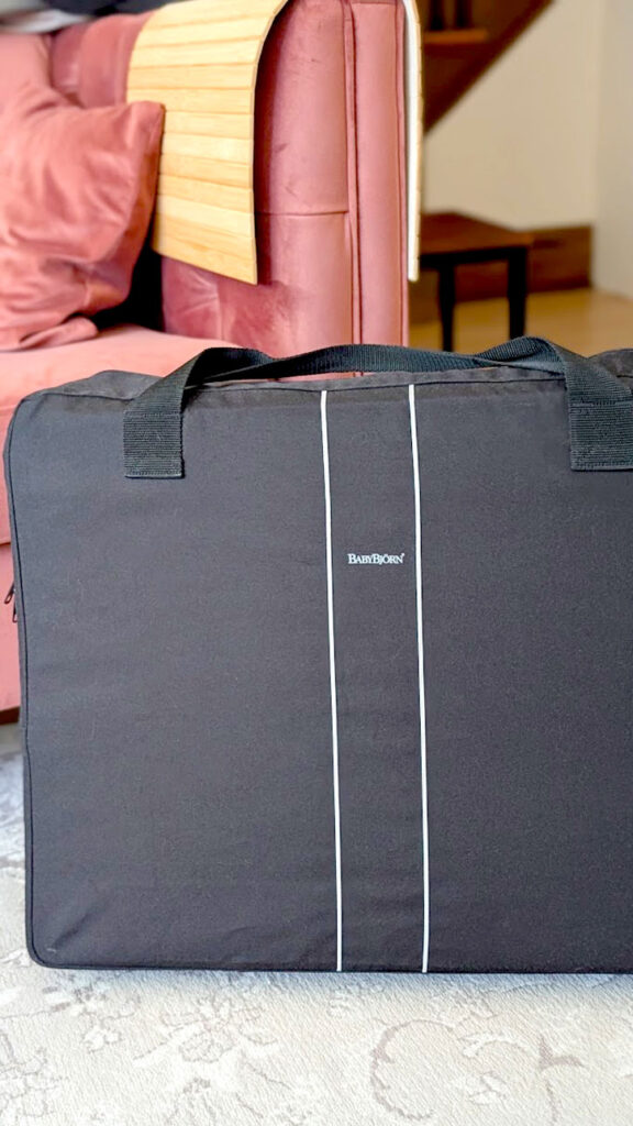 the babybjörn travel case sits on the floor in the house, in front of a pink couch. It's a black carry bag, that is rectangular, looking like a large briefcase carry bag. 