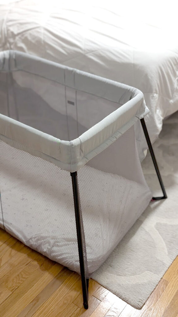 the babybjörn travel crib is sit up ready to be used in a bedroom with a bed behind it. The legs are black, which doesn't match the grey fabric of the crib.