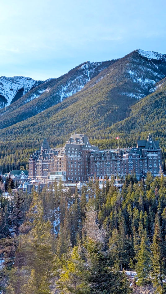 a brown chateau hidden amongst the trees of the mountains in the Canadian rockies, there's a bit of snow on the top of the mountains.