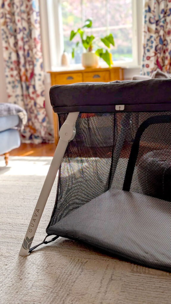 The guava lotus travel crib where you can clearly see the white leg sticking out holding up the travel crib frame. On the white leg, it says Guava with their logo. The bed sits in a beautifully sunny room, with fun patterned draped opening up to let the light in through the window. 
