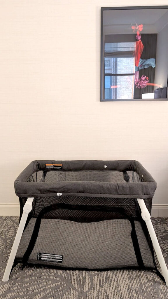 directly in the middle of the photo, very plane is the guava lotus travel crib. The legs are white and stick out, while the crib is made out of grey mesh. The wall behind it is pure white, making the travel crib pop.