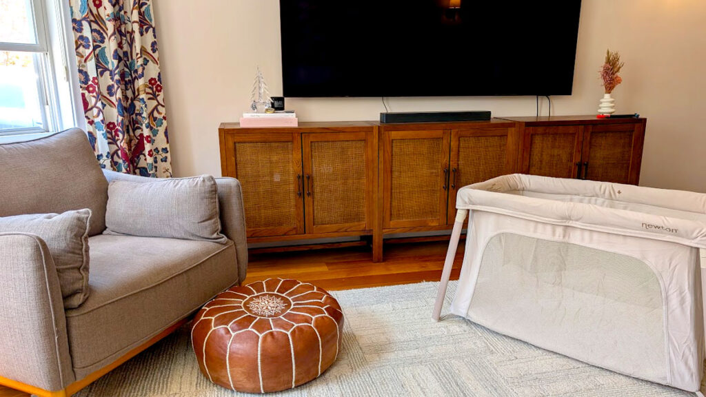 the newton compact travel crib sits on the floor in the den, with a large tv behind it and an armchair with a poof directly in front of it, fitting right in to the house.