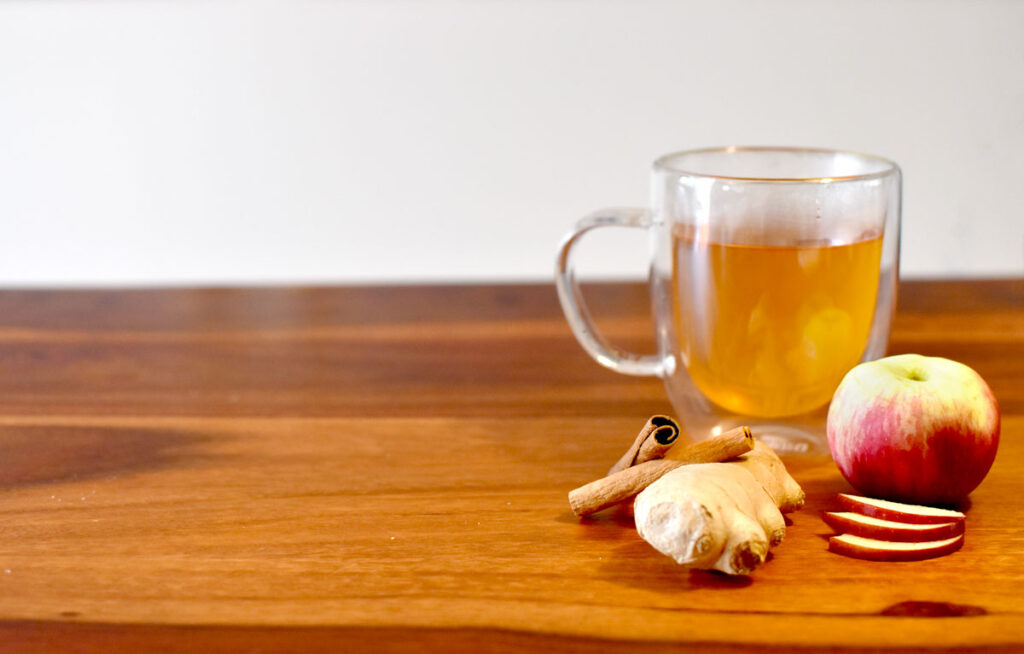 one a wooden table sits a doubled walled glass mug with an orange liqueur cinnamon apple tea. In front of it sits all the main ingredients, an apple, cinnamon sticks, and ginger