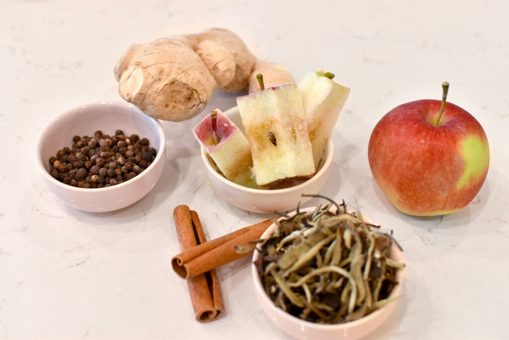 5 apple tea ingredients on the counter, a full gingerroot in the back. three pinch bowls, one is filled with peppercorn, one with three frozen apple cores, and in front is a pile of white tea leaves. There are also two cinnamon sticks on the counter and a full apple for decor.
